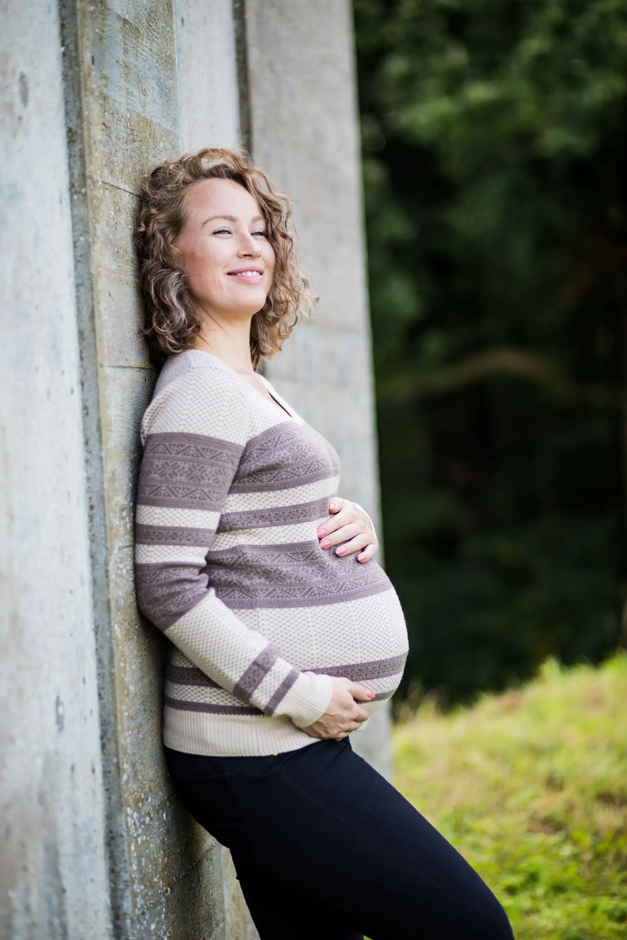 Smilende gravid kvinde i stribet strikbluse læner sig op ad en mur med hænderne om maven