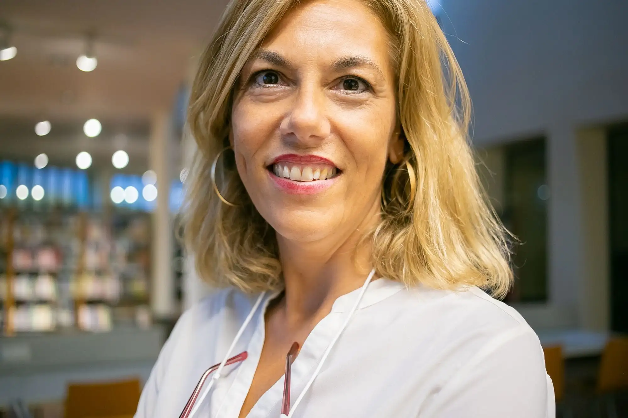 Smilende kvinde med blondt hår og store øreringe i hvid bluse i et bibliotek