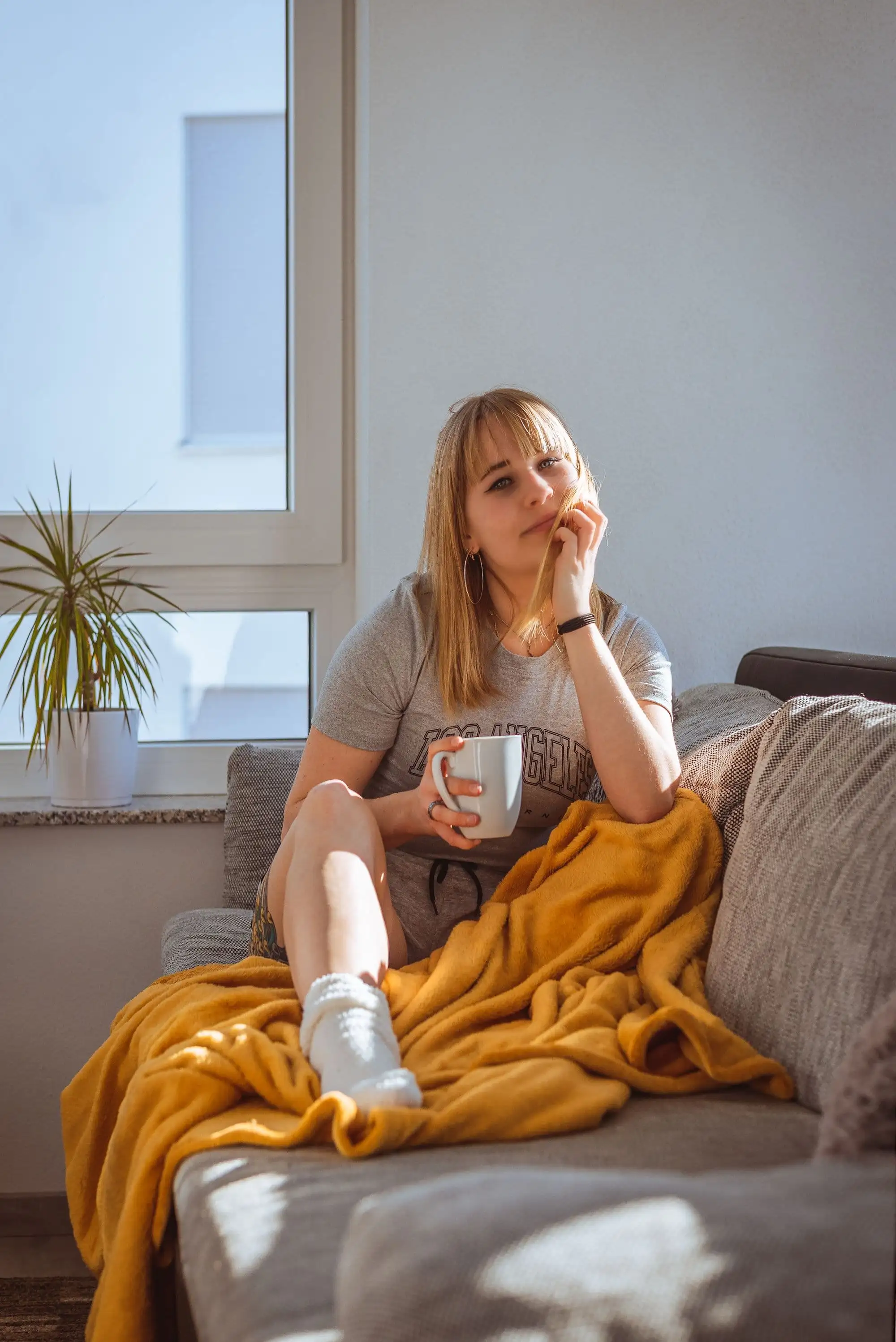 Kvinde i grå T-shirt sidder afslappet i en sofa med et gult tæppe og et krus