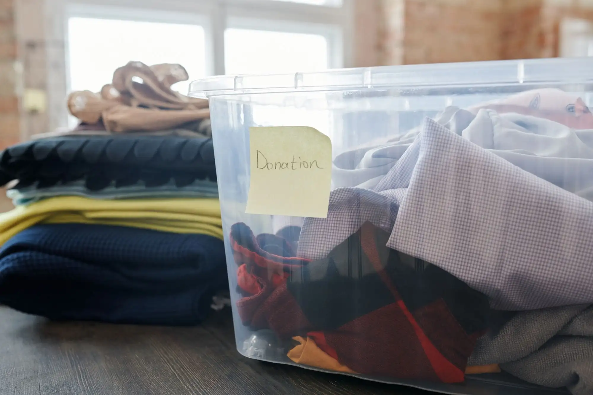 Gennemsigtig plastkasse med brugt tøj og en seddel med ordet Donation ved siden af stakke af sammenfoldet tøj